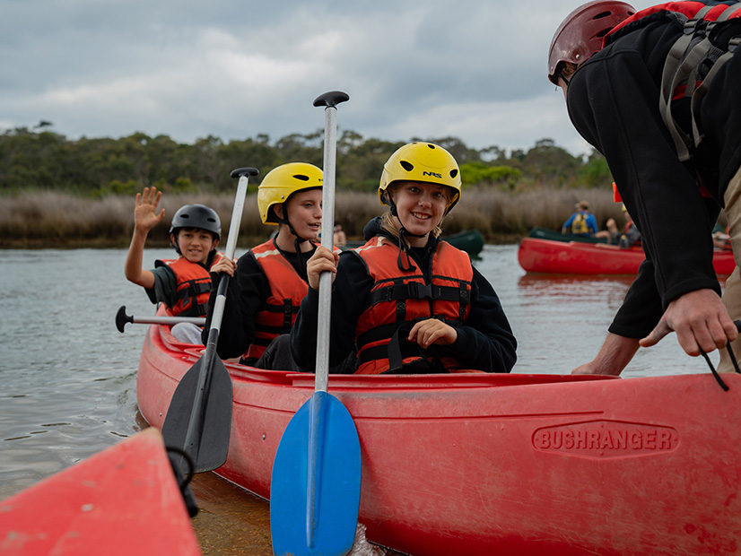 Three students sat in a red canoe wearing helmets and life jackets, while holding paddles and waving