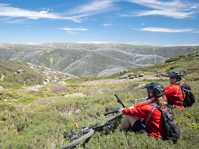 Two mountain bike riders are sitting at the summit of Bogong High Plains which overlooks Falls Creek Village.