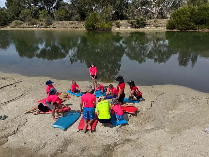 An outdoor scene featuring a group of children and adults gathered on a sandy riverbank next to calm water. Most children wear bright pink or red long-sleeved rash vests, dark shorts, and wide-brimmed hats, sitting or lying on blue and red bodyboards. A yellow-shirted instructor with 'INSTRUCTOR' written on the back kneels at the center, addressing the group. The background showcases a tranquil body of water reflecting lush green trees from the opposite bank, creating a serene natural setting.