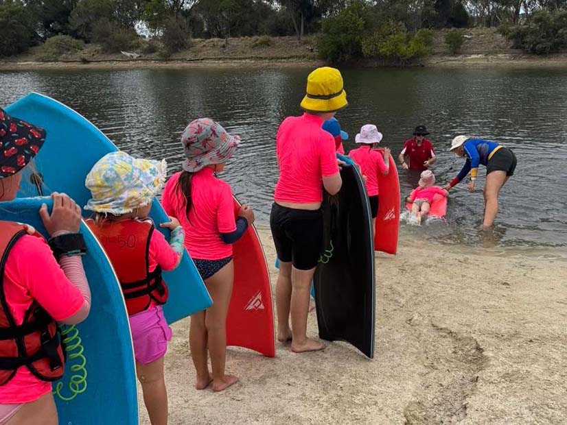 A vibrant outdoor scene at a calm body of water, featuring children and adults engaged in water activities. In the foreground, several children stand on a sandy bank holding boogie boards, wearing bright pink long-sleeved tops, bucket hats, and some with orange life vests. In the mid-ground, adults supervise a baby in a pink inflatable floatation device in the shallow water. The background showcases lush green trees lining the far bank, creating a natural and serene setting